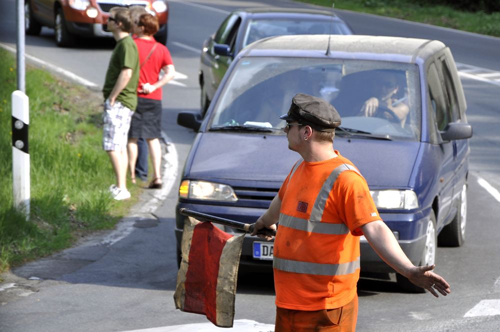 Ein Mann in orangenfarbener Uniform hält den PKW Verkehr auf und sichert den Bahnuebergang